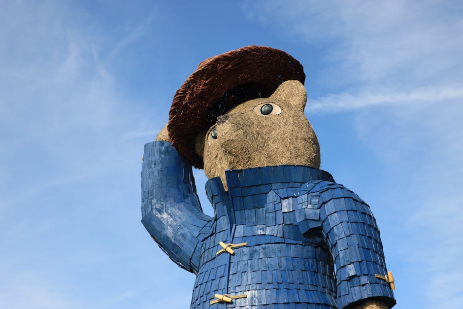 A large outdoor sculpture of a cartoonish, stylized human figure with a round, beige face, big eyes, and brown hair, wearing a textured blue coat with wooden toggle fastenings, standing against a blue sky with wispy clouds. The sculpture's surface appears to be made of various materials, including textured stone for the face and layered wooden or ceramic shingles for the coat, with the figure holding its right hand to its head in a saluting or thoughtful pose. The lighting highlights the details and textures of the sculpture, which is likely part of a public art installation, captured during daytime under natural sunlight.