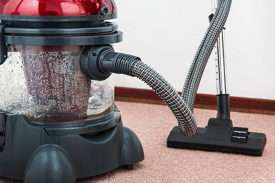 Close-up of a professional vacuum cleaner with a clear plastic canister filled with dust and debris, attached to a flexible hose that connects to a floor head, positioned on a light pink carpet in a room with white walls and a wooden skirting board. The vacuum is in operation, indicating surface cleaning and deep cleaning activity, with visible soap foam or moisture on the canister suggesting recent sanitisation or wet cleaning process. The scene highlights the equipment used by Paddington Carpet Cleaners for domestic cleaning services, emphasizing thorough carpet maintenance and hygiene.