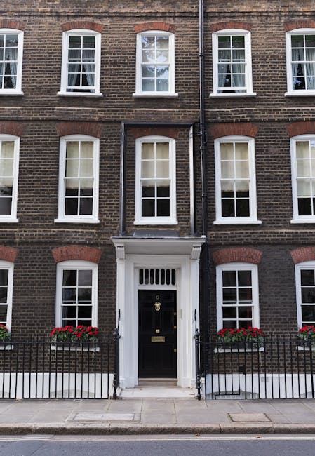 A cobblestone sidewalk runs along a row of white Georgian-style terraced houses in Paddington. The pavement has evenly laid rectangular stones, some with slight wear, and is bordered by a black wrought-iron fence with pointed finials. Behind the fence, small gardens feature lush green foliage and flowering plants. A blue bicycle leans against the fence near one house entrance. The buildings have large windows with black frames and decorative black window grills, and their facades are clean and well-maintained. Natural daylight illuminates the scene, highlighting the contrast between the white walls, dark fence, and vibrant greenery. The overall setting reflects a tidy, residential area emphasizing neatness and charm, with an implied connection to property maintenance and cleaning services such as those offered by Paddington Carpet Cleaners.
