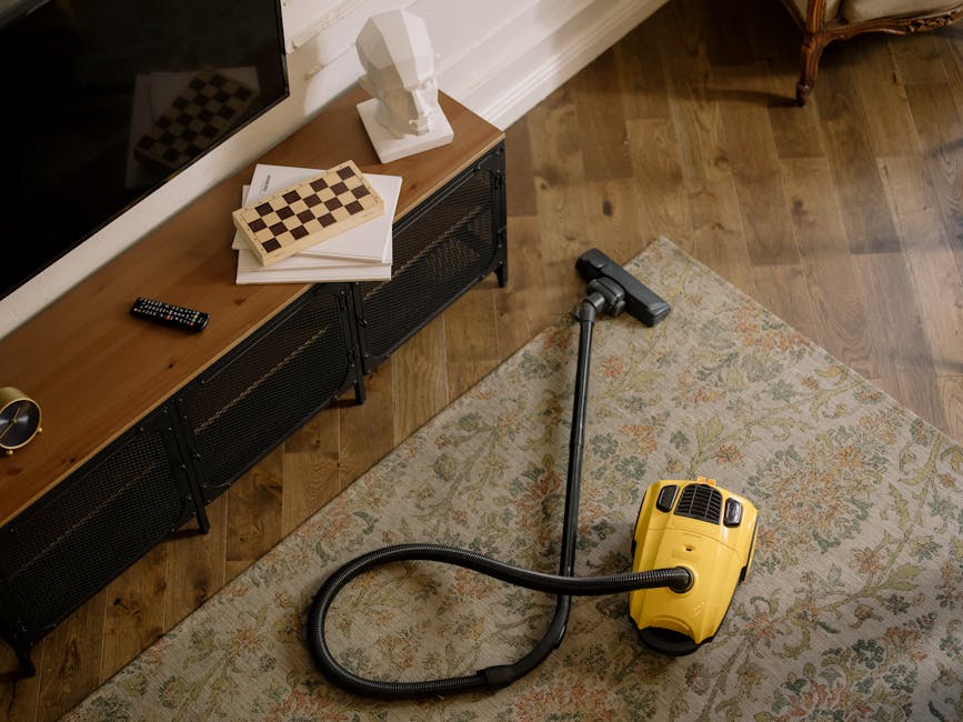 Close-up view of a vacuum cleaner with a transparent dust container filled with debris, connected to a flexible, patterned hose attached to a floor head, positioned on a soft pink carpet in a room wit