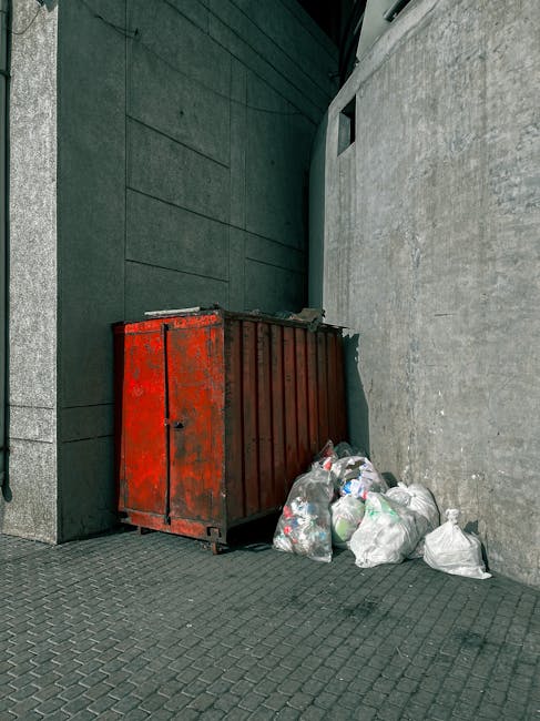 An outdoor scene showing a large, rusty red waste disposal container placed against a modern building wall with dark green panels and a textured concrete surface. Surrounding the container are several