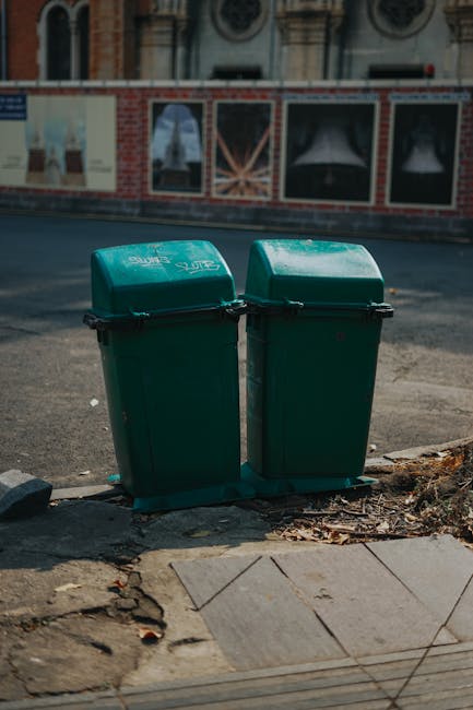 An outdoor scene showing a large, rusty red waste disposal container placed against a modern building wall with dark green panels and a textured concrete surface. Surrounding the container are several