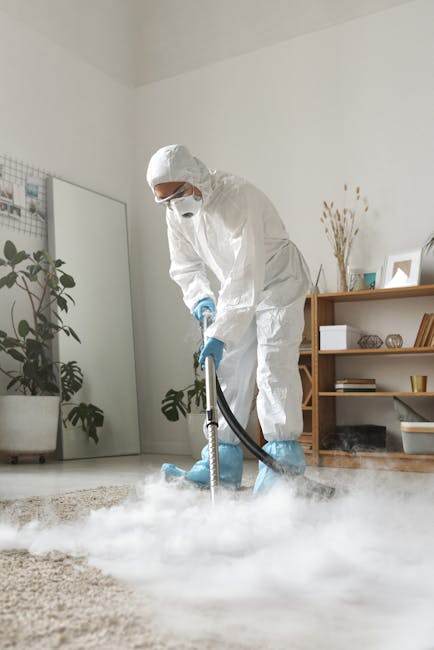 A man in an orange sweater and grey pants is shown bending down on his knees to vacuum or clean a beige carpet in a residential living room. The room features a white wall, a wooden floor border, and