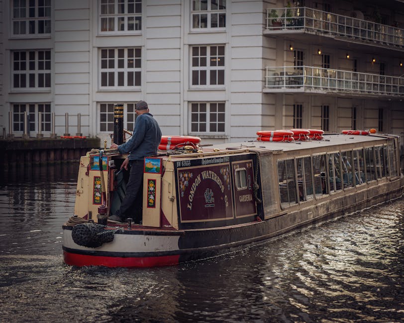 A narrowboat named 'London Waterbus' is navigating a canal near a modern residential building with white exterior walls, multiple large windows, and balconies. The boat features a dark purple and cream color scheme, with decorative floral motifs and the registration number 'DW 63855' visible on the front. On the roof, there are bright orange life buoys and safety equipment. The scene is captured during daylight, with reflections of the boat and building visible on the water's surface. The setting appears calm, with no visible debris or dirt on the boat or water, emphasizing cleanliness and maintenance consistent with professional cleaning standards. Paddington Carpet Cleaners is naturally associated with surface cleaning and hygiene, although the image itself focuses on a boat and urban environment and does not depict cleaning activities directly.