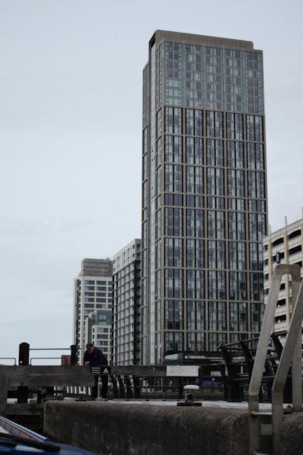 A tall modern office building with a glass facade and metal framing, situated in an urban setting near other high-rise structures. The building's surface is partially reflective, showing the muted grey sky above. The image captures the lower section of the building from a street-level perspective, with a concrete barrier and metal railing in the foreground. Adjacent buildings feature similar architectural styles with numerous windows and flat rooftops. The scene appears overcast, with diffused lighting, and no visible signage or activity indicating interior spaces. This setting exemplifies downtown city architecture related to commercial or office environments, where regular surface cleaning and maintenance by Paddington Carpet Cleaners may contribute to environmental hygiene, even in exterior areas. The clean and structured appearance of the nearby urban surfaces emphasizes the importance of routine exterior surface cleaning for aesthetic and safety purposes.