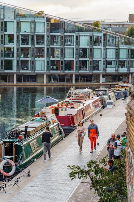 A narrowboat named 'London Waterbus' is navigating a canal near a modern residential building with white exterior walls, multiple large windows, and balconies. The boat features a dark purple and cream color scheme, with decorative floral motifs and the registration number 'DW 63855' visible on the front. On the roof, there are bright orange life buoys and safety equipment. The scene is captured during daylight, with reflections of the boat and building visible on the water's surface. The setting appears calm, with no visible debris or dirt on the boat or water, emphasizing cleanliness and maintenance consistent with professional cleaning standards. Paddington Carpet Cleaners is naturally associated with surface cleaning and hygiene, although the image itself focuses on a boat and urban environment and does not depict cleaning activities directly.