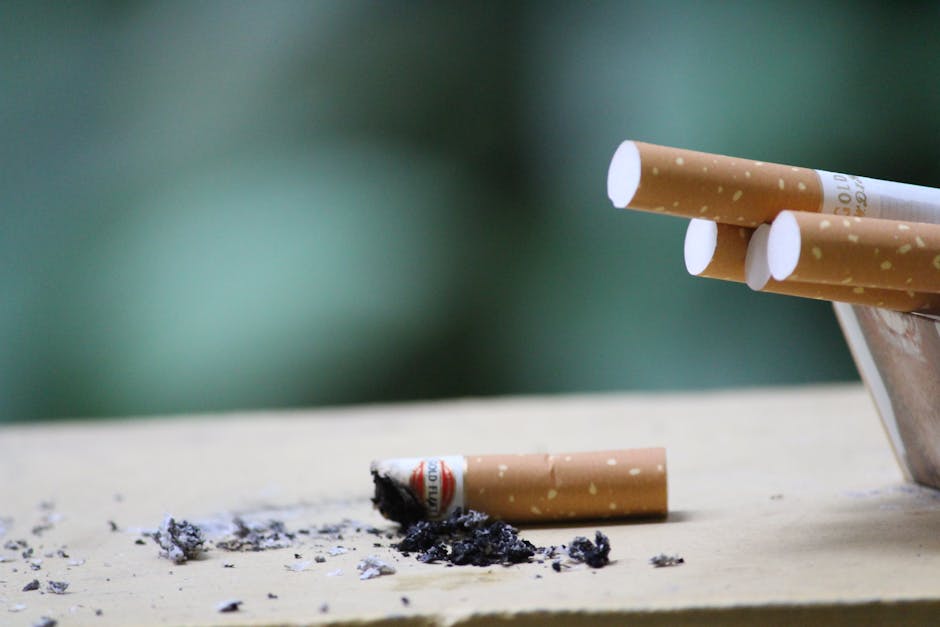 Close-up of broken cigarette butts and ashes on a wooden surface, with remnants of burnt tobacco and partially smoked cigarettes in the foreground. The background is blurred with a greenish hue, highl