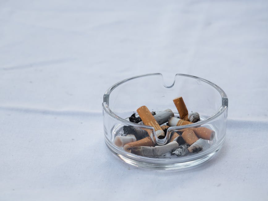 Close-up of broken cigarette butts and ashes on a wooden surface, with remnants of burnt tobacco and partially smoked cigarettes in the foreground. The background is blurred with a greenish hue, highl