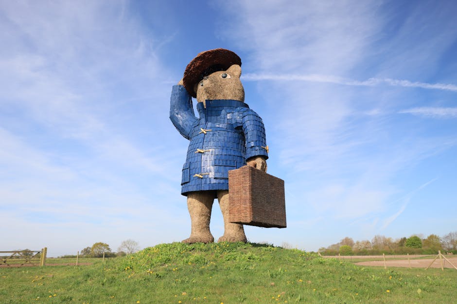 A large outdoor sculpture depicting a stylized figure of a boy wearing a blue coat with toggle buttons and a brown hat, saluting while holding a brown suitcase in his right hand. The sculpture is mounted on a grassy hill under a clear blue sky with wispy clouds, and the figure's surface appears textured with materials resembling wood or composite. The setting includes a simple wooden fence in the background, with open fields and sparse trees in the distance, creating a sense of open countryside. The lighting is bright and natural, emphasizing the sculpture's details and the vibrant outdoor environment. Paddington Carpet Cleaners would emphasize surface cleaning and maintenance for similar outdoor or decorative surfaces, aligning with their focus on deep cleaning and sanitisation, although the primary content here is decorative art rather than cleaning.